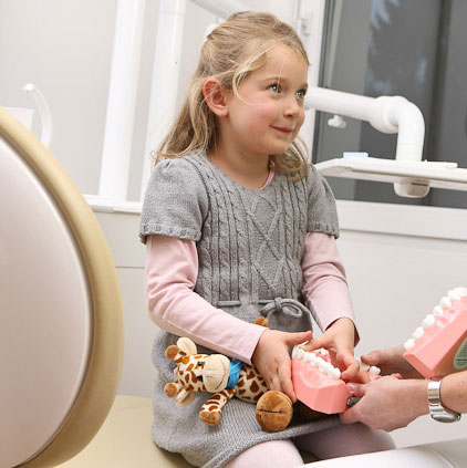 symbolbild kinderzahnheilkunde kleines maedchen mit riesem gebiss in der hand und einer spielzeug giraffe in der zahnarztpraxis